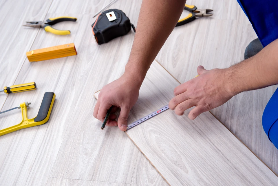 Artisan en bleu travaillant à genoux sur parquet stratifié clair, mesurant une lame avec mètre ruban et crayon