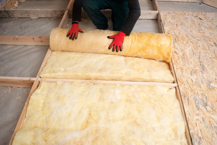 Ouvrier installant de l'isolant thermique entre les solives d'un plancher, portant des gants de protection rouges