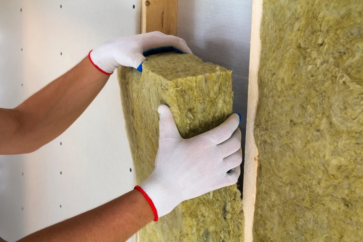 Intérieur de bâtiment en construction, ouvrier en combinaison de protection blanche et gants installant de la laine de verre jaune entre montants métalliques d'une cloison