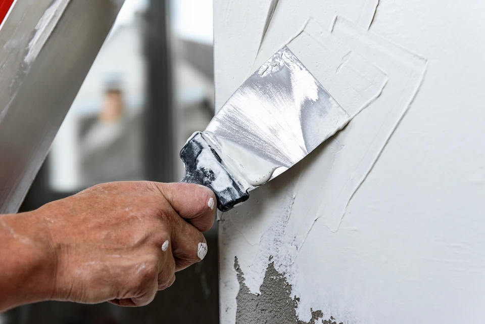Scène intérieure de travaux de finition où une main applique un enduit ou mortier sur une surface de béton brut à l'aide d'une taloche rectangulaire